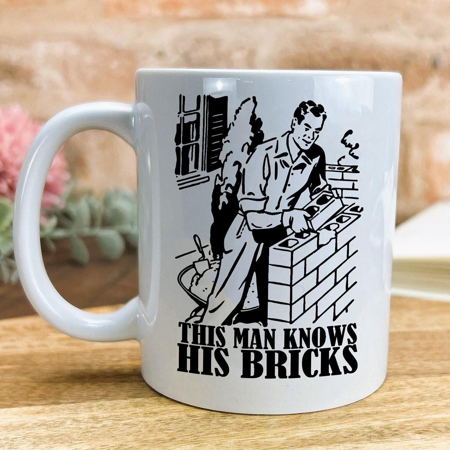 A white ceramic mug with a black and white illustration of a man working on bricks, along with the text "This man knows his bricks" printed on it.