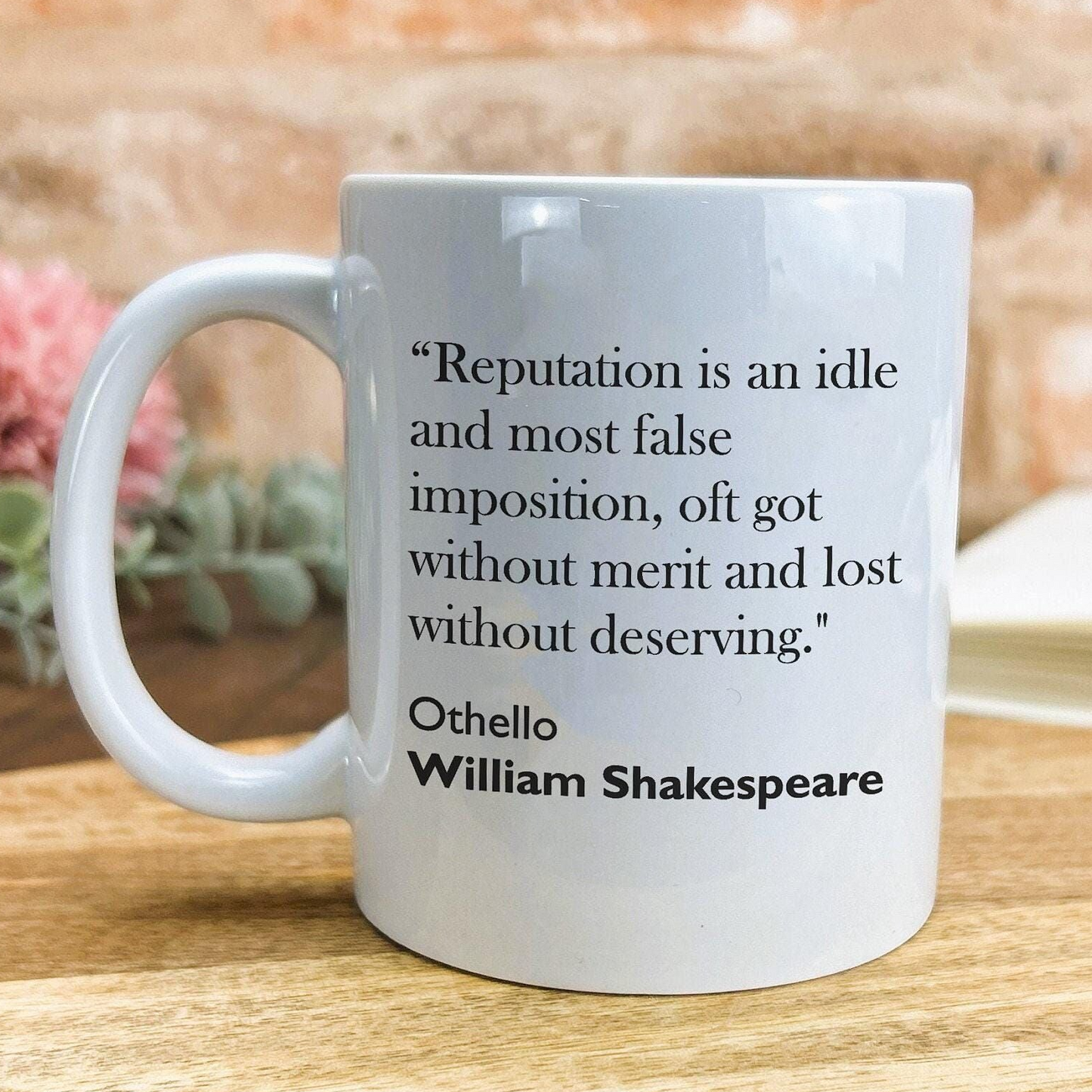 A white ceramic mug with a quote from William Shakespeare printed on it, placed on a wooden surface.
