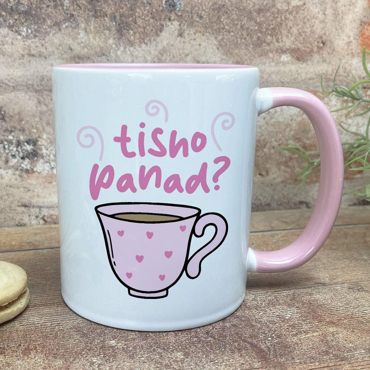 A white mug with a pink handle, featuring a cartoon illustration of a teacup with hearts on it, and the text "tisho panad?" written above it.