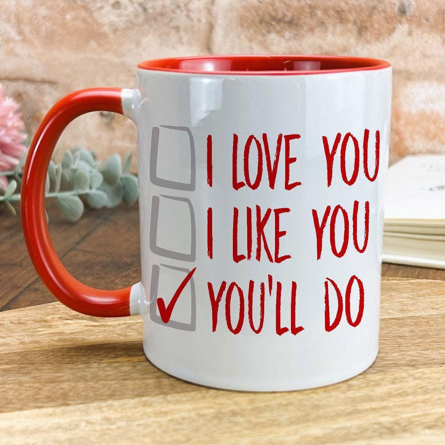 A white mug with a red handle, featuring a humorous text that reads "I LOVE YOU I LIKE YOU YOU'LL DO" along with checkboxes.