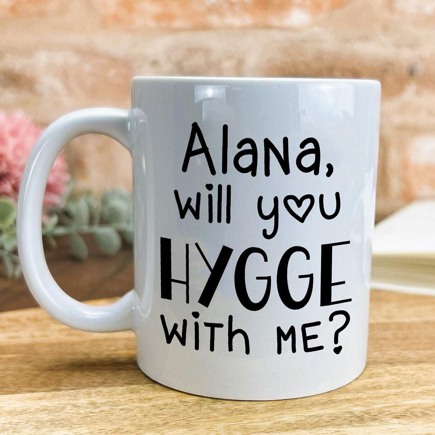 A white ceramic mug with black text that reads "Alana, will you hygge with me?"