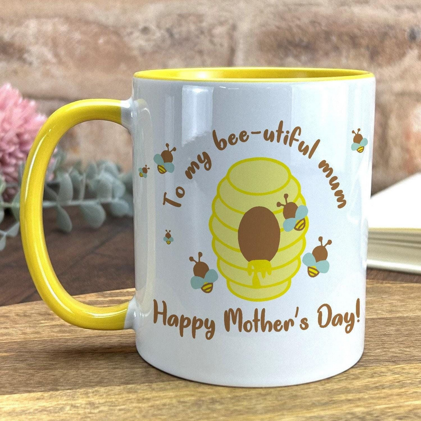 A white mug with a yellow rim, featuring a bee and honeycomb design, along with the text "To my bee-utiful mum Happy Mother's Day!"