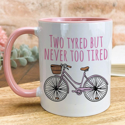 A white mug with a pink handle, featuring a bicycle illustration and the text "Two Tyred But Never Too Tired" printed on it.