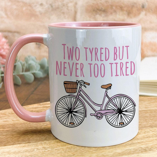 A white mug with a pink handle, featuring a bicycle illustration and the text "Two Tyred But Never Too Tired" printed on it.