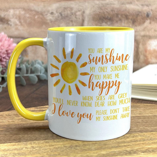 A white coffee mug with a yellow handle, featuring a sun design and text that reads "You are my sunshine, my only sunshine, you make me happy. When skies are grey, dear how much I love you. Please, don't take my sunshine away".