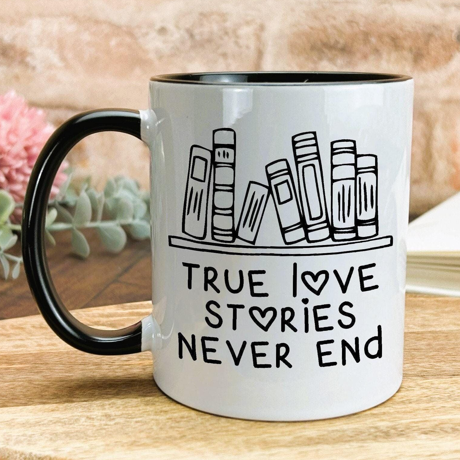 A white mug with a black handle, featuring a graphic design of books and the text "True / Love / Stories / Never End" printed on it.