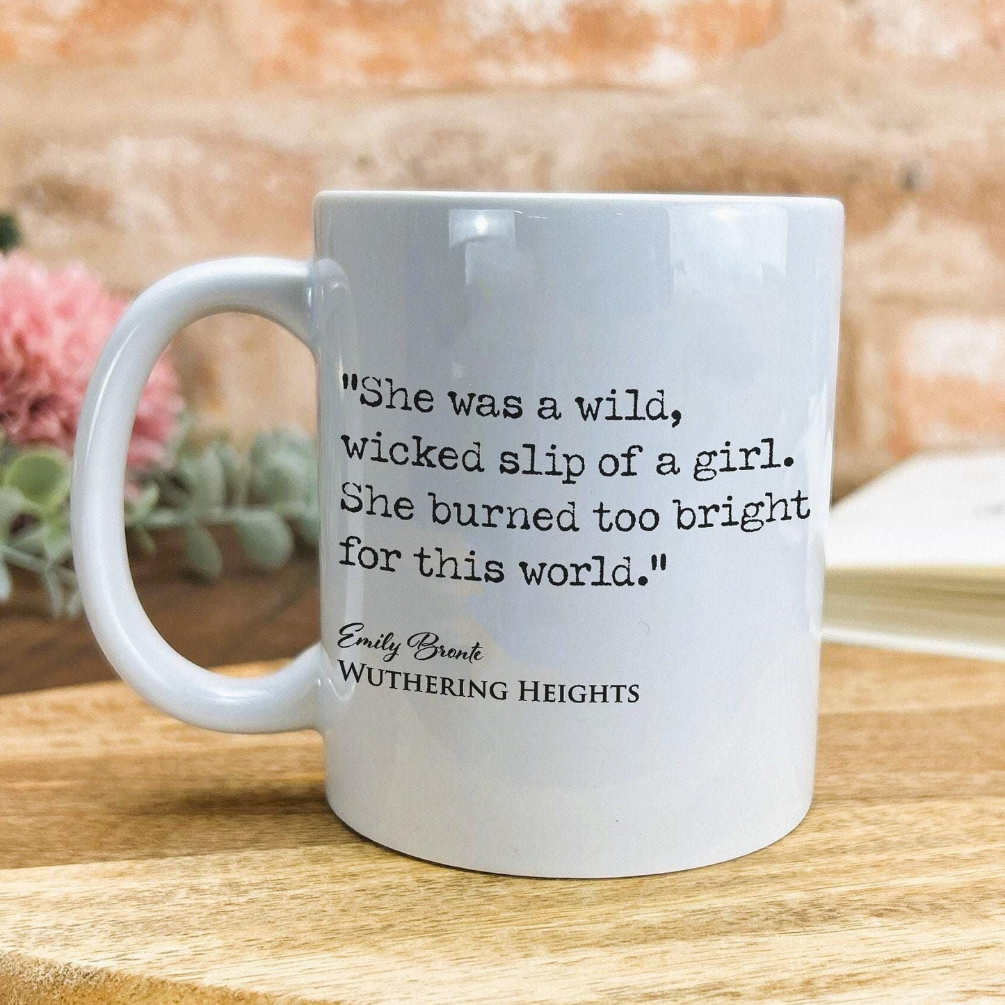 A white ceramic mug with a quote from the novel "Wuthering Heights" printed on it, placed on a wooden surface against a brick wall background.