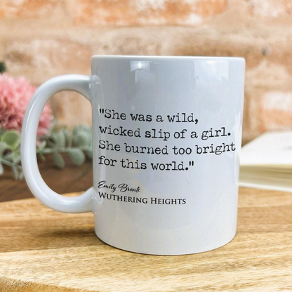 A white ceramic mug with a quote from the novel "Wuthering Heights" printed on it, placed on a wooden surface against a brick wall background.