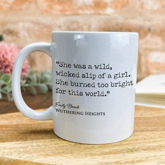 A white ceramic mug with a quote from the novel "Wuthering Heights" printed on it, placed on a wooden surface against a brick wall background.