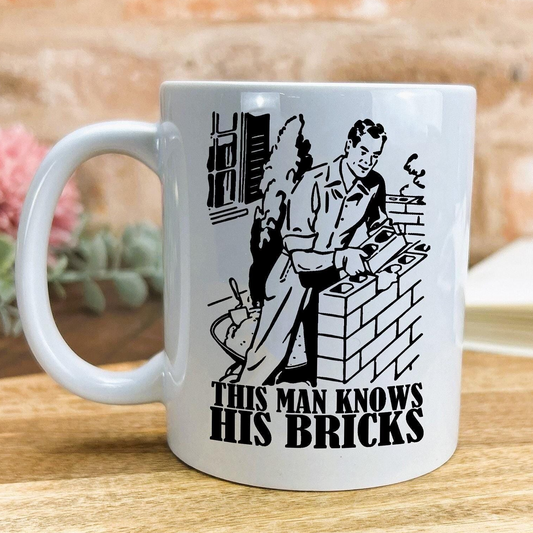 A white ceramic mug with a black and white illustration of a man working on bricks, along with the text "This man knows his bricks" printed on it.