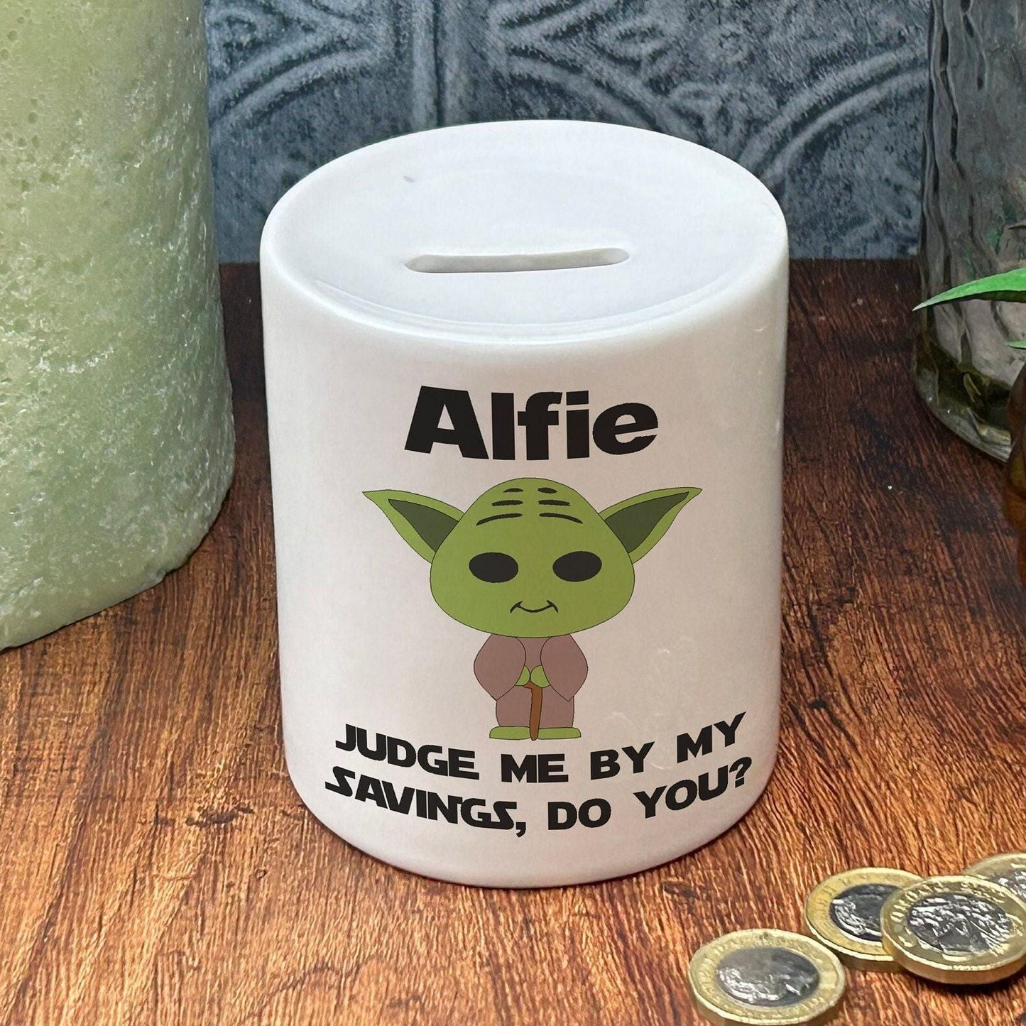 A white ceramic coin bank with a cartoon image of a green alien character and text that says "Alfie" and " Judge me by my savings, do you?".