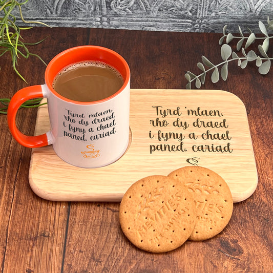 A mug of coffee and two cookies are placed on a wooden tray, accompanied by a plant and a wooden cutting board with a message in Welsh.