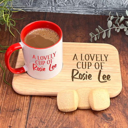 A red and white mug with the text "A Lovely Cup of Rosie Lee" sits on a wooden cutting board, accompanied by two cookies and some greenery.