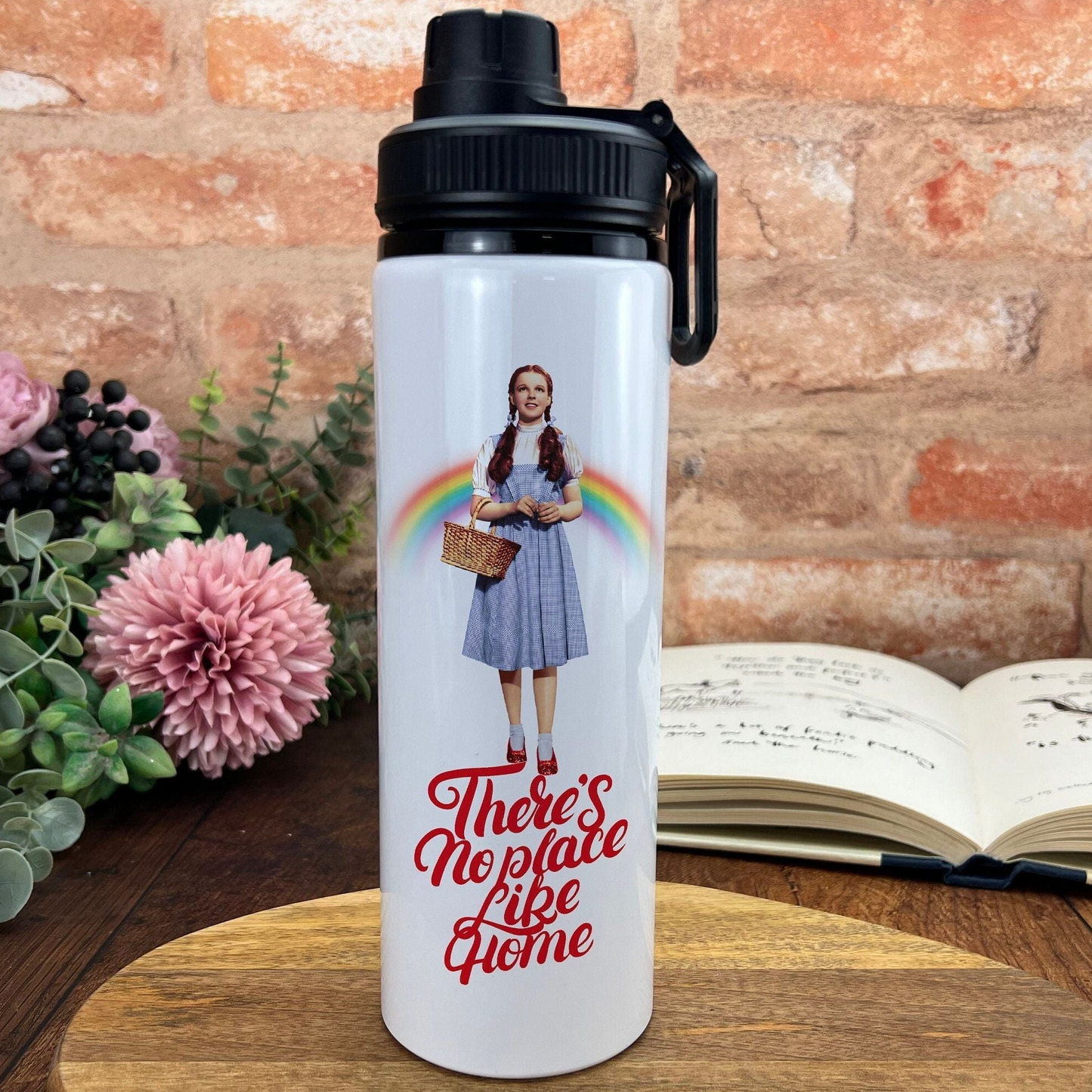 A white water bottle with a printed image of a woman and the text "There's No Place Like Home" on it, placed on a wooden surface next to a book and some flowers.