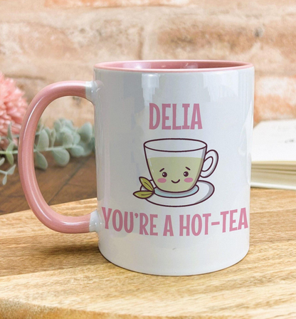 A white mug with a pink handle, featuring a cartoon cup and the text "DELIA YOU'RE A HOT-TEA" printed on it.