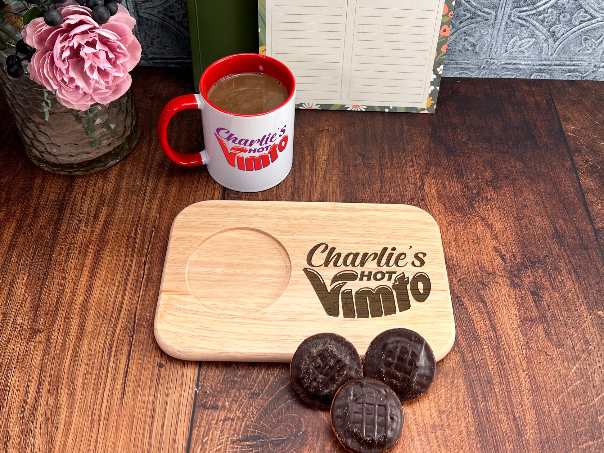 A wooden tray with a coffee mug and a note, along with a vase of pink flowers and a book, all placed on a wooden surface.