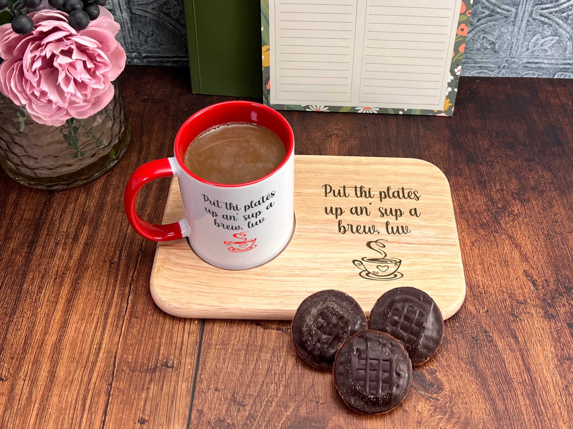 A wooden tray with a message about putting plates up for a brew, accompanied by a mug of coffee and three chocolate-covered cookies on a wooden table.