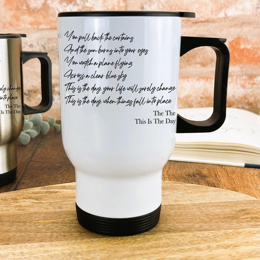 A white travel mug with black text and a black handle, placed on a wooden surface.