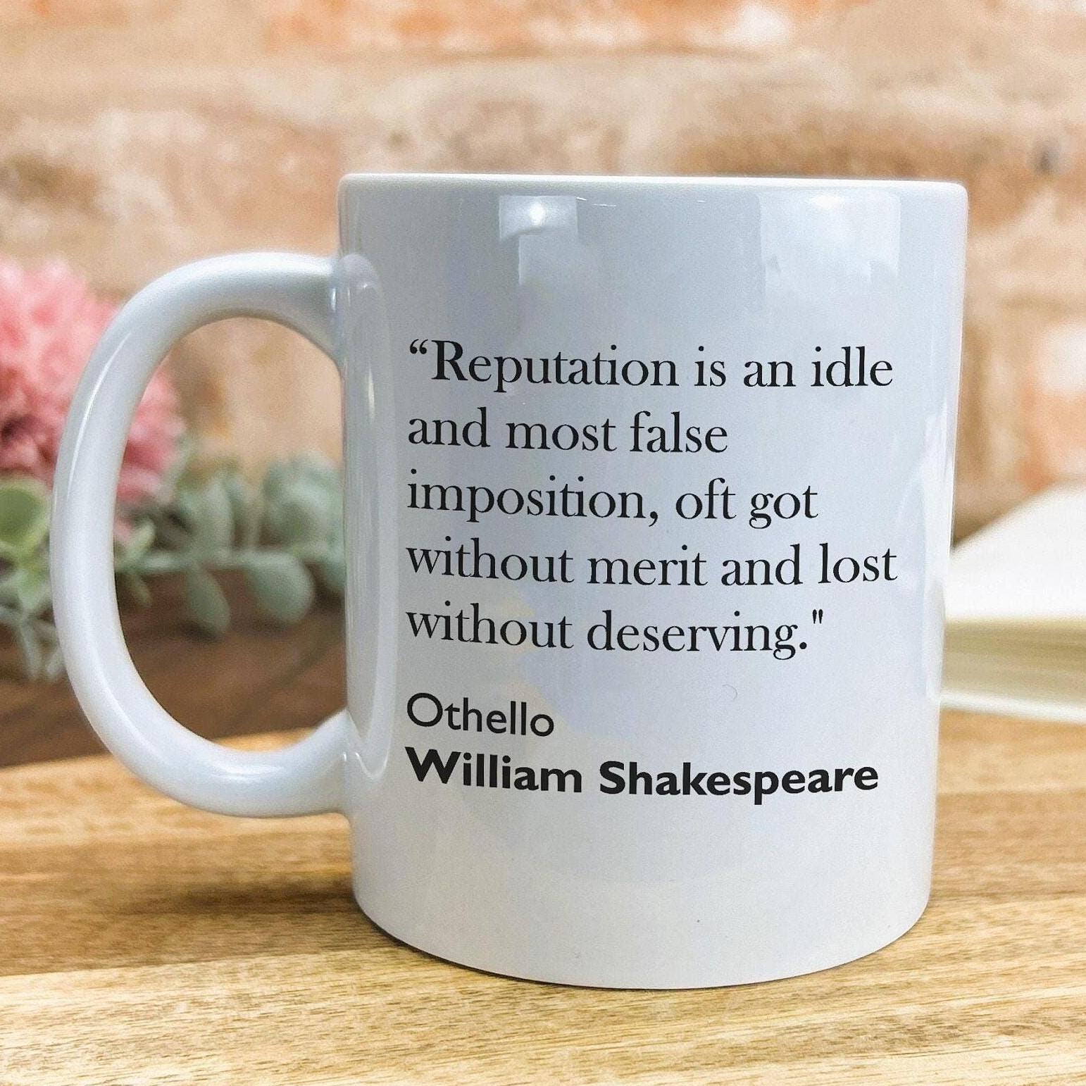 A white ceramic mug with a quote from William Shakespeare printed on it, placed on a wooden surface.