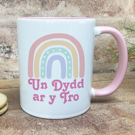 A white mug with a pink handle, featuring a colorful rainbow design and the Welsh phrase "Un Dydd ar y Tro" (which means "Happy Day" in English).