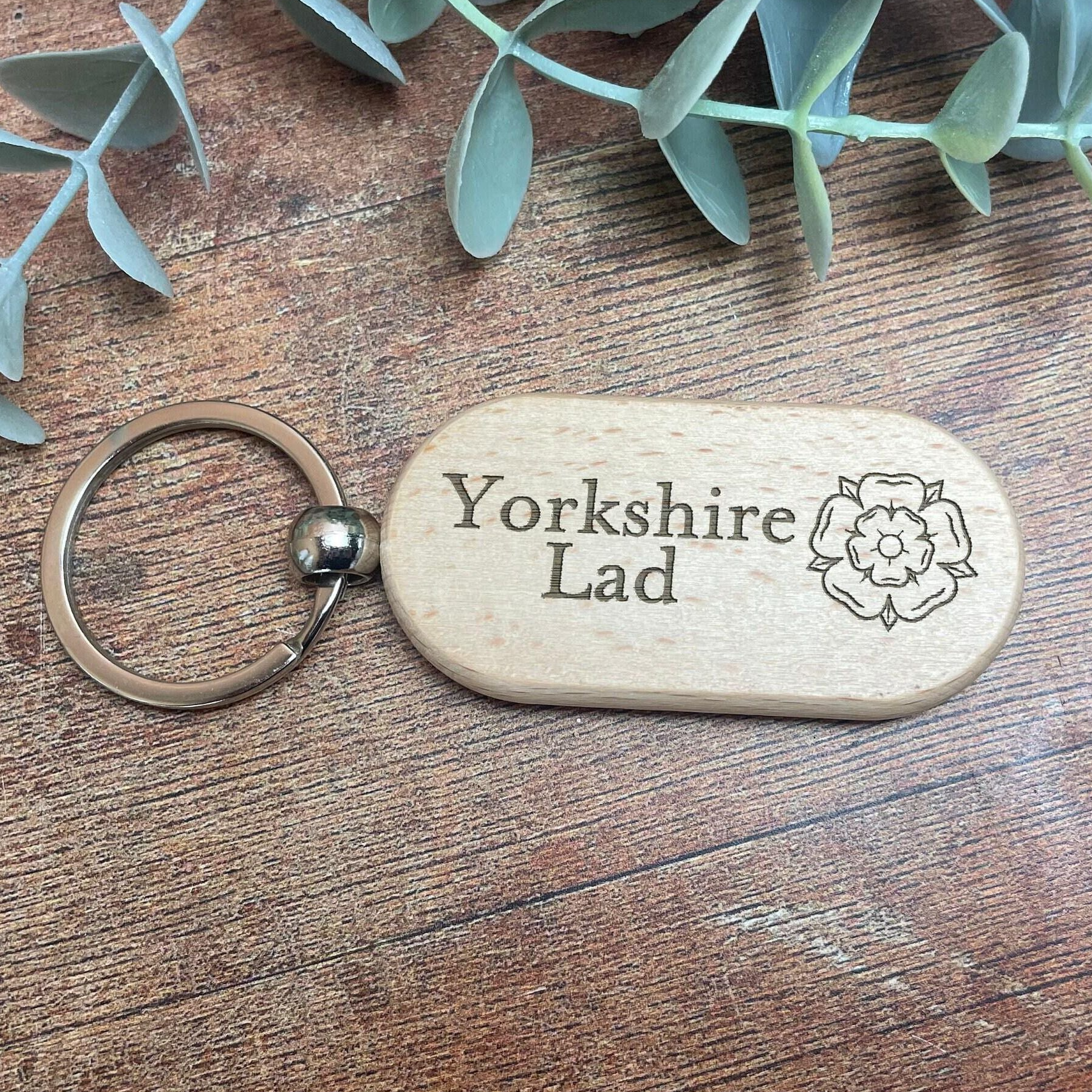 A wooden keychain with the text "Yorkshire Lad" engraved on it, placed on a wooden surface with some greenery in the background.