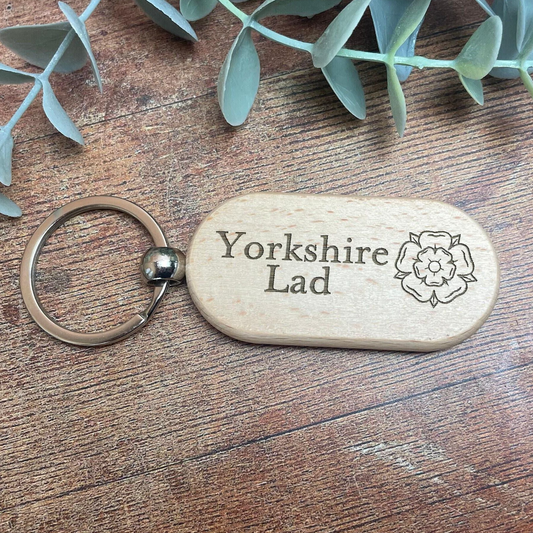 A wooden keychain with the text "Yorkshire Lad" engraved on it, placed on a wooden surface with some greenery in the background.
