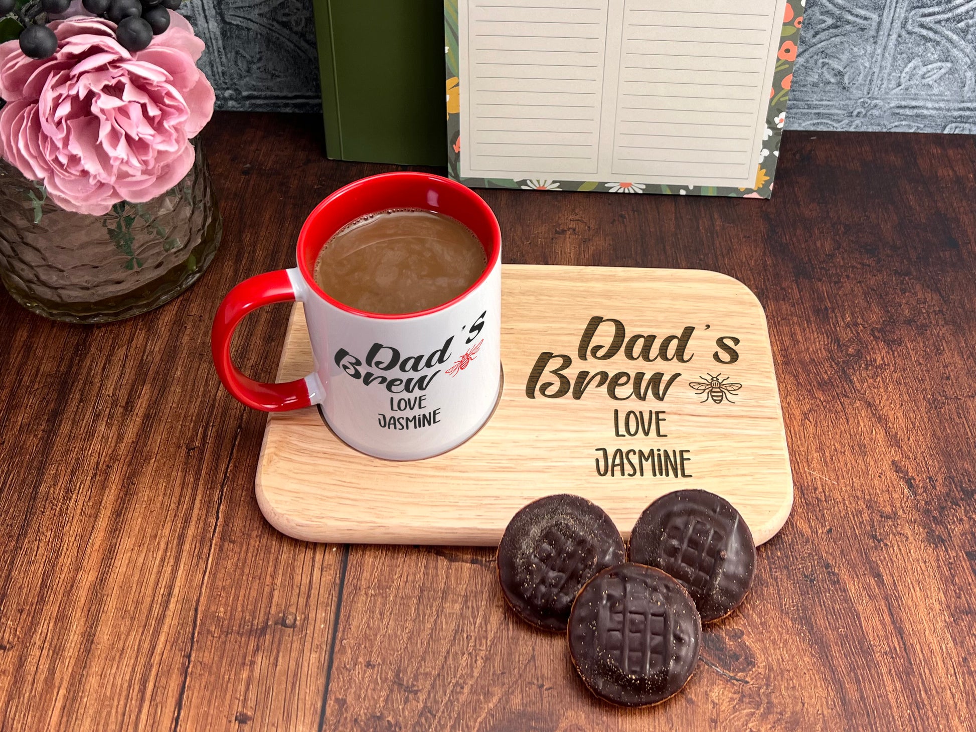A red and white mug labeled "DAD'S BREW" sits on a wooden tray, accompanied by three chocolate-covered cookies and a vase of pink flowers.