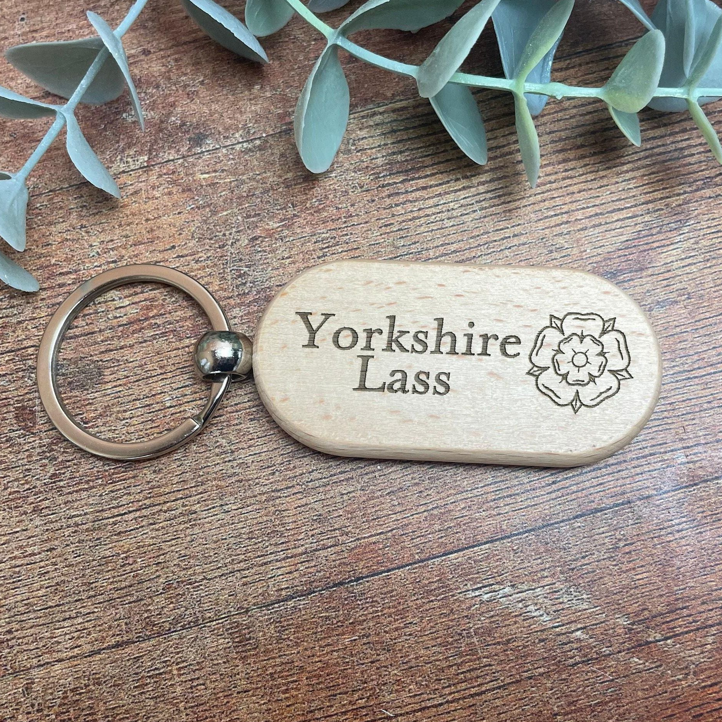 A wooden keychain with the text "Yorkshire Lass" engraved on it, placed on a wooden surface with some greenery in the background.