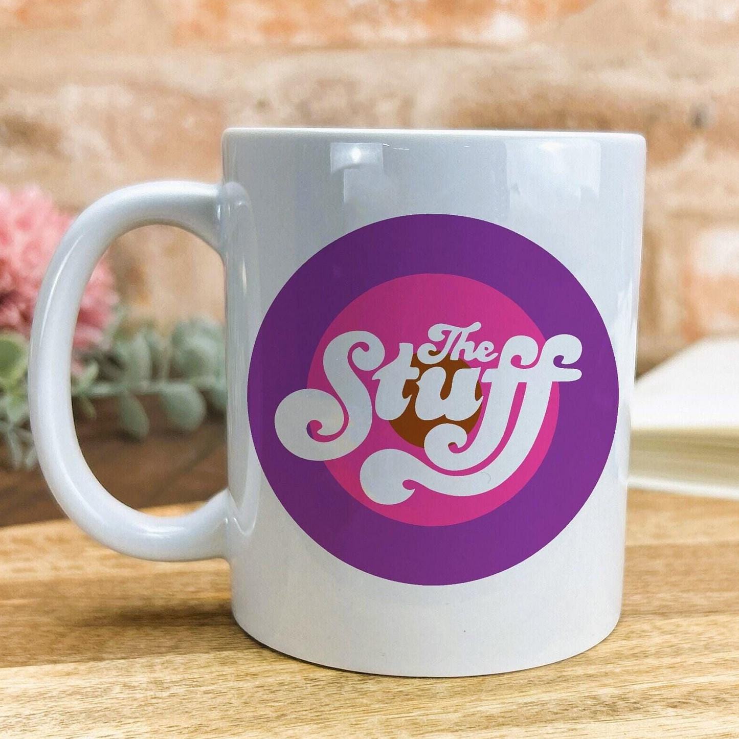 A white ceramic mug with a purple and pink circular logo featuring the text "The Stuff" on it.