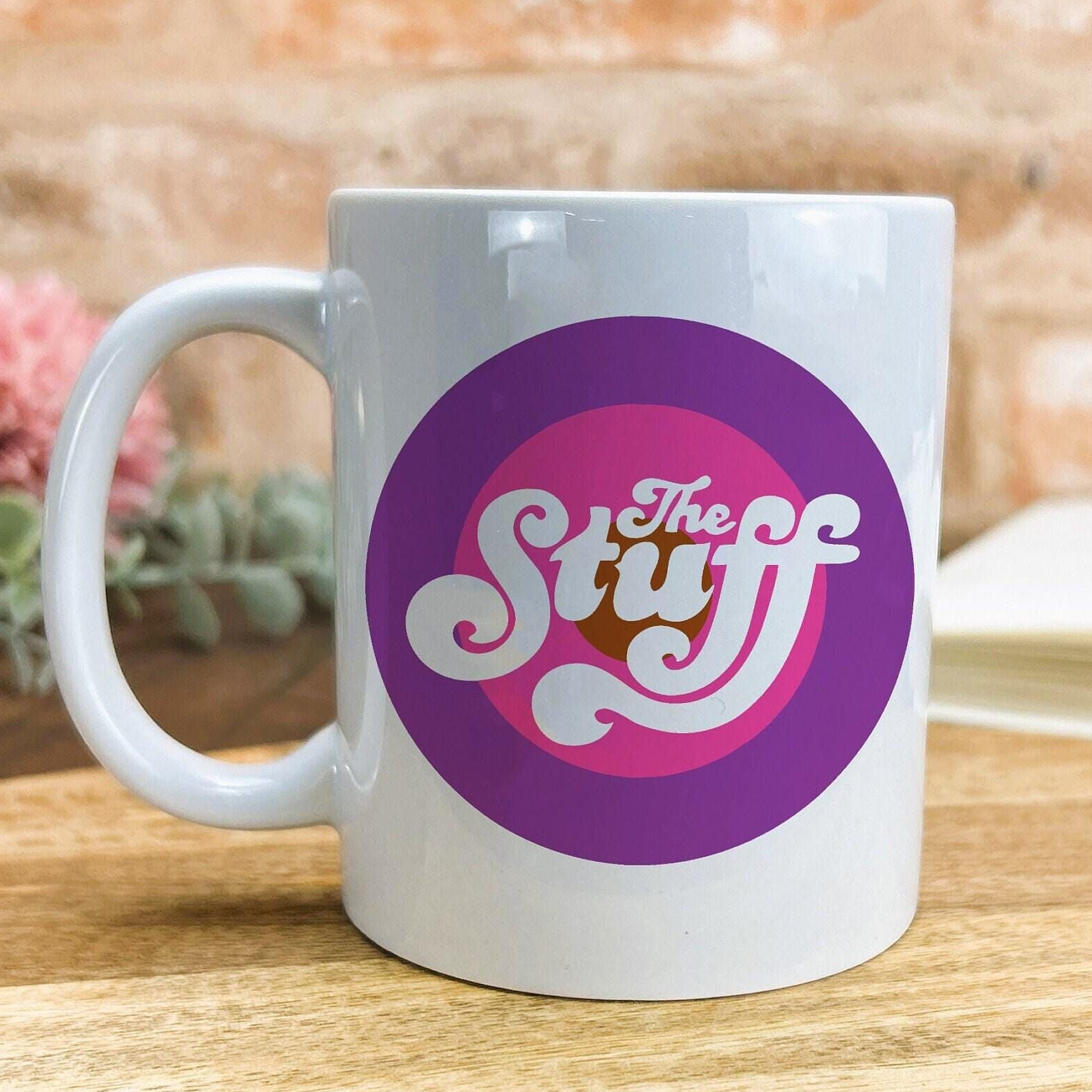 A white ceramic mug with a purple and pink circular logo featuring the text "The Stuff" on it.