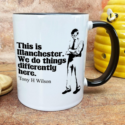 A white mug with a black handle and a black and white illustration of a man with glasses. The mug has text that reads "This is Manchester. We do things differently here. Tony H Wilson".