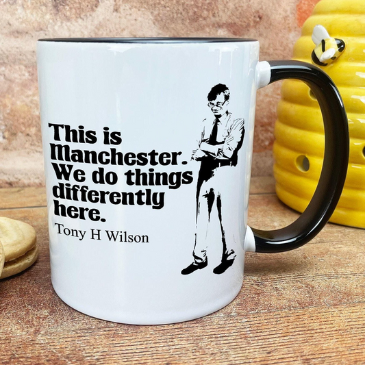 A white mug with a black handle and a black and white illustration of a man with glasses. The mug has text that reads "This is Manchester. We do things differently here. Tony H Wilson".