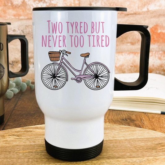 A white travel mug with a black handle and lid, featuring a pink bicycle illustration and the text "Two Tyred But Never Too Tired" printed on it.