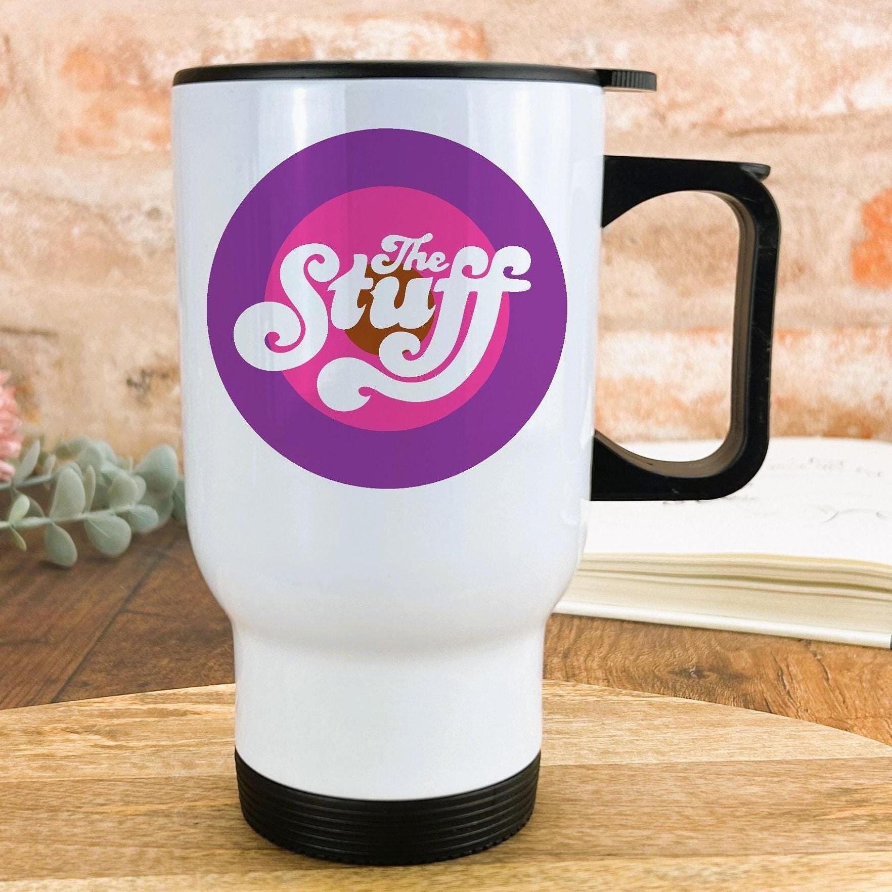 A white travel mug with a black handle, featuring a purple and pink circular logo with the text "The Stuff" printed on it.