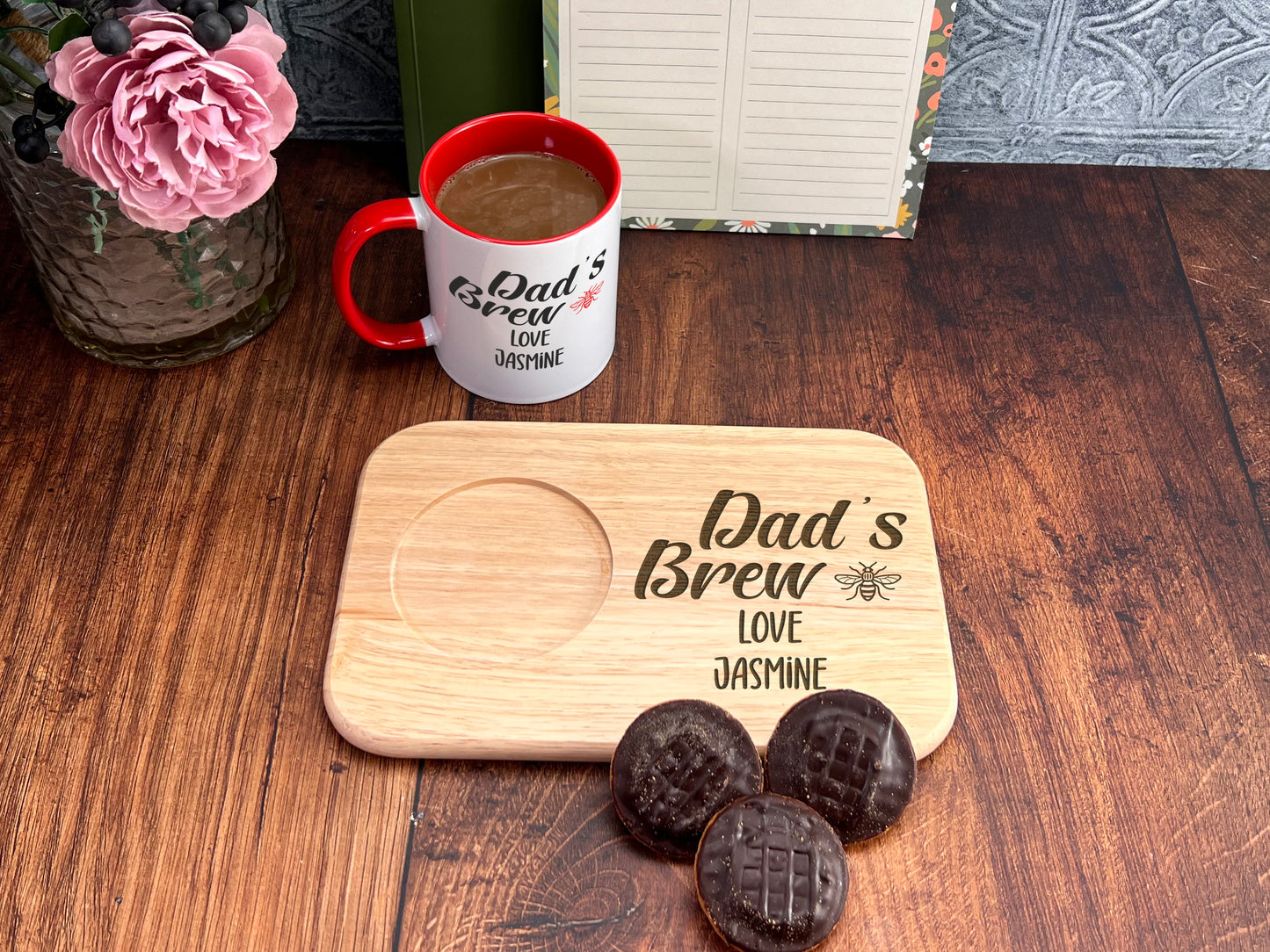 A wooden tray with a message that says "Dad's Brew" and "Love Jasmine" is placed on a wooden table, accompanied by a mug and a vase with pink flowers. There are also chocolate cookies and a book on the table.