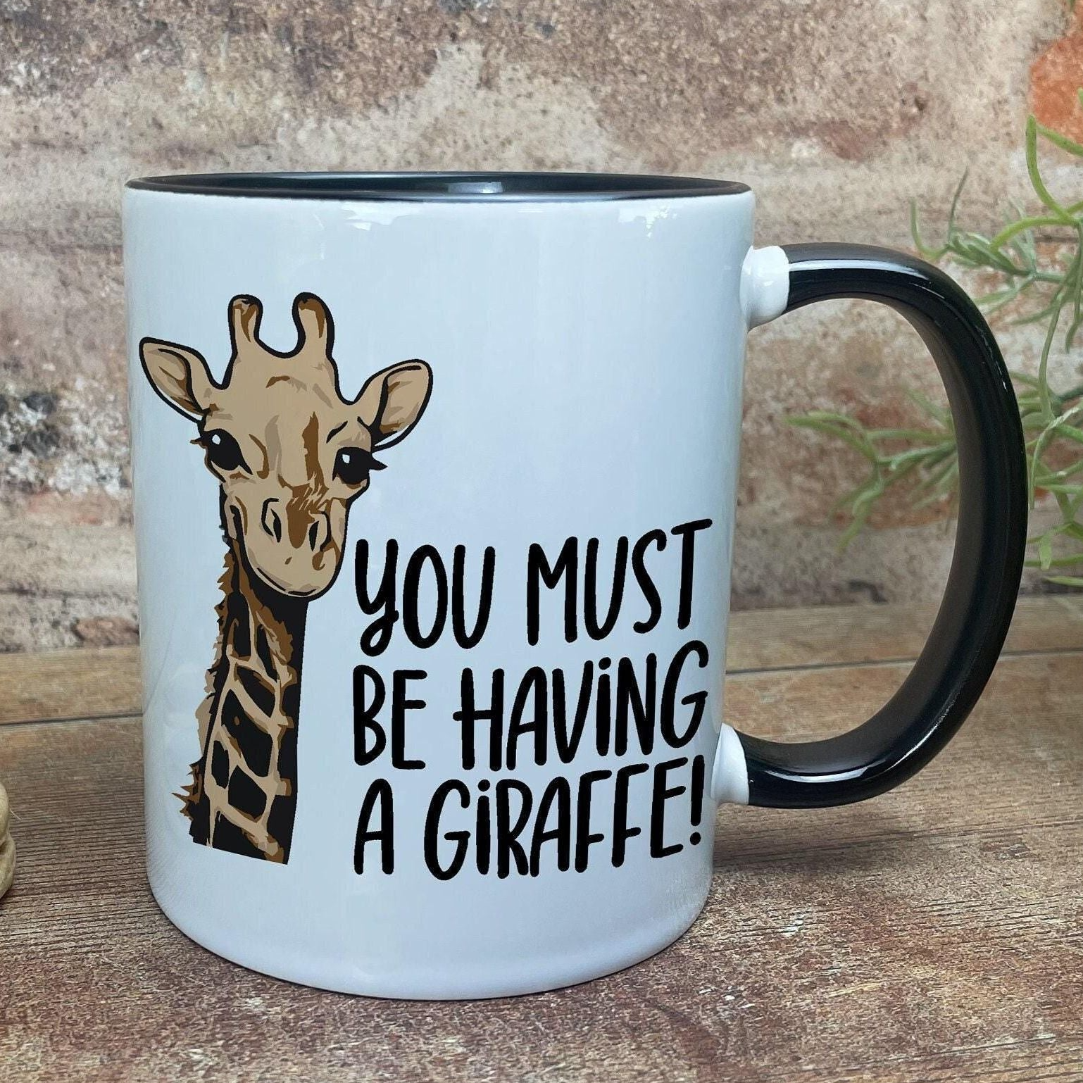 A white mug with a black handle, featuring an illustration of a giraffe and the text "You MUST BE HAVING A GIRaffe!"