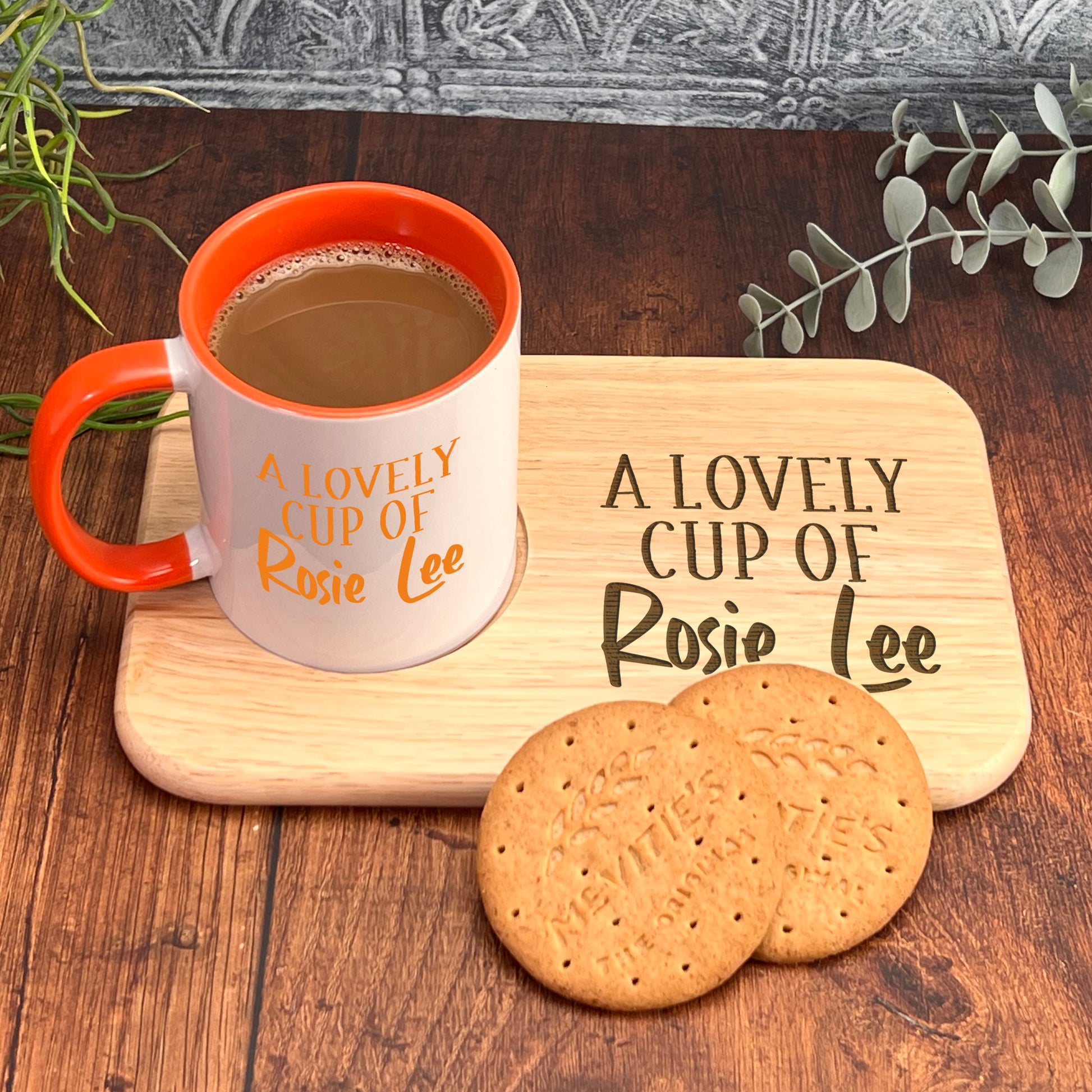 A white mug with an orange rim and the text "A LOVELY CUP OF Rosie Lee" sits on a wooden tray, accompanied by two round cookies.