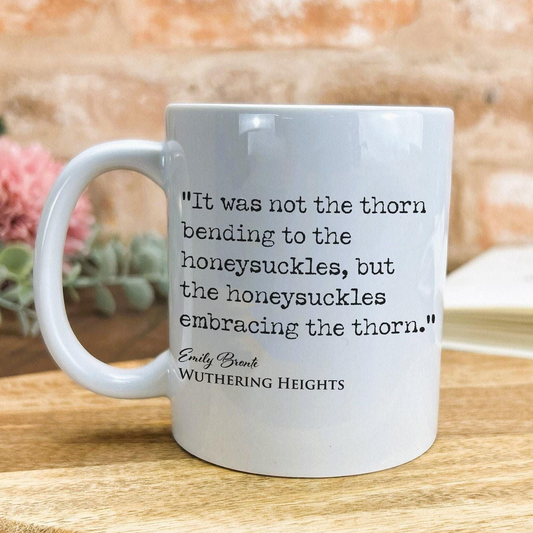 A white ceramic mug with a quote from the novel "Wuthering Heights" by Emily Brontë printed on it, placed on a wooden surface against a brick wall background.