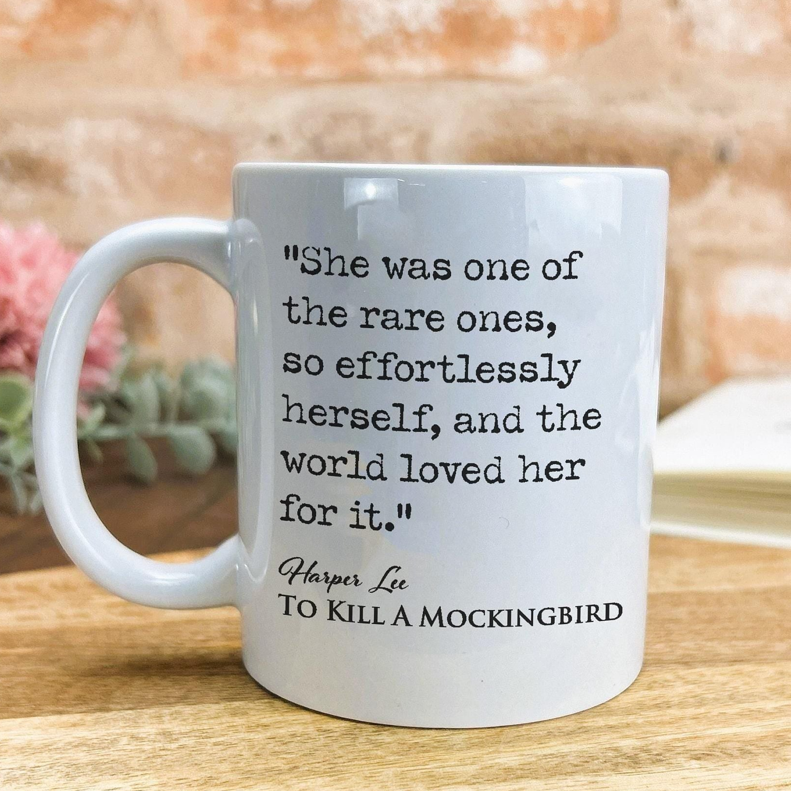 A white ceramic mug with a quote from the novel "To Kill a Mockingbird" printed on it, placed on a wooden surface against a brick wall background.