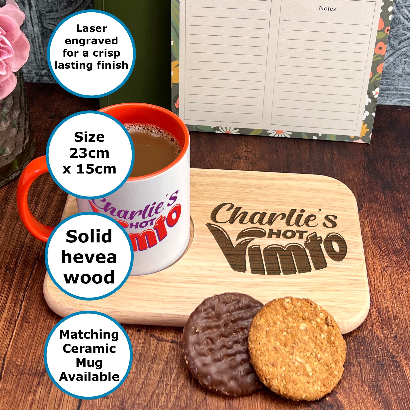 A wooden tray with a mug and two cookies, accompanied by a note pad and a vase of flowers, on a wooden table.