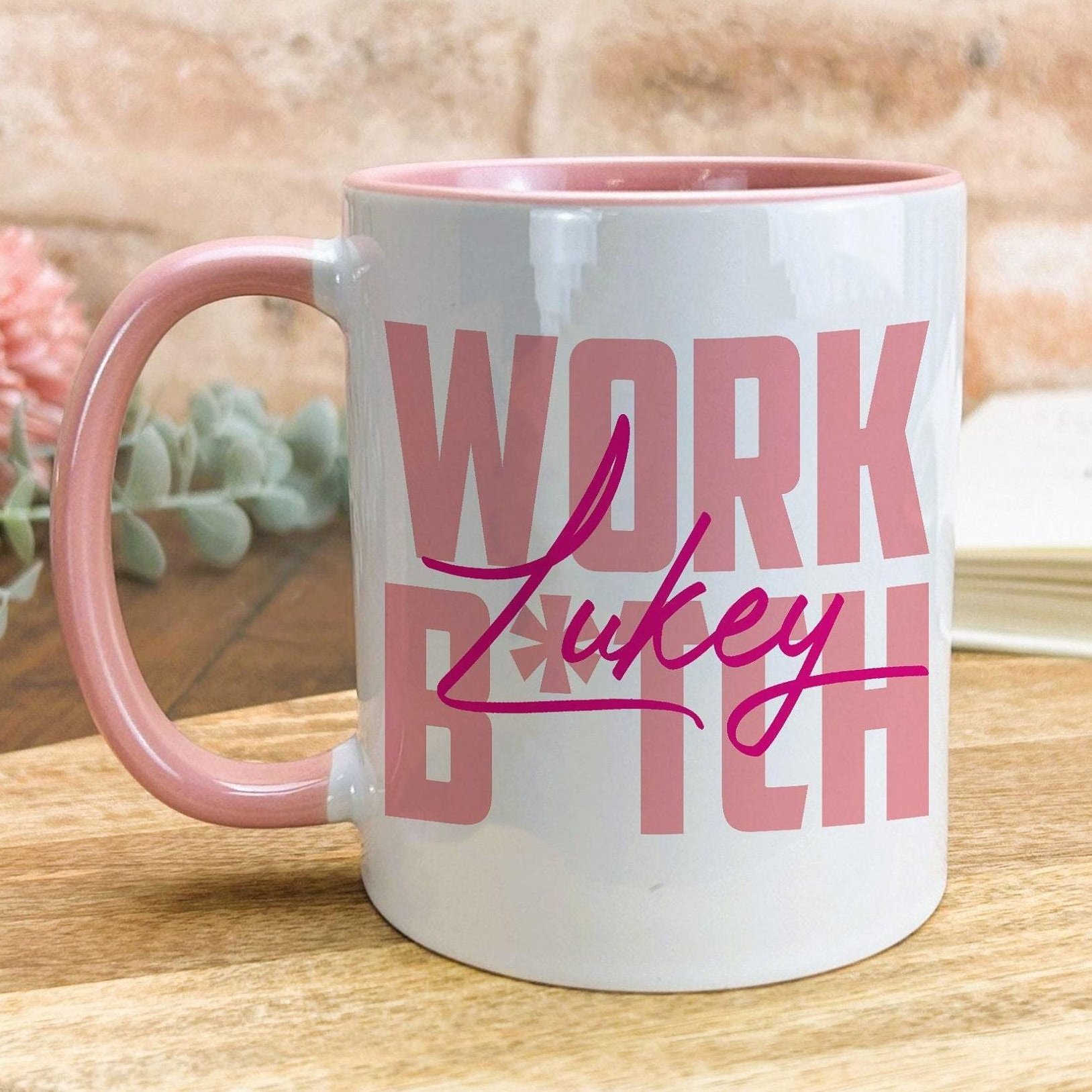 A white mug with a pink handle, featuring the text "WORK LUCKY" in pink letters.