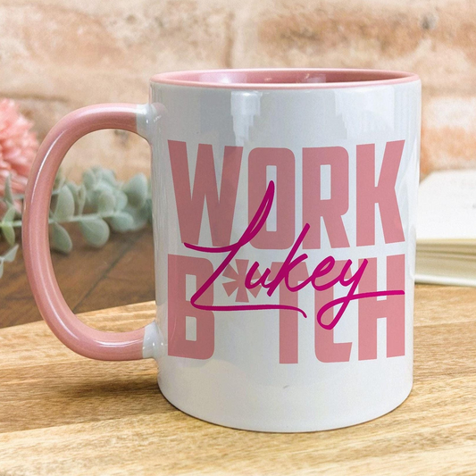 A white mug with a pink handle, featuring the text "WORK LUCKY" in pink letters.