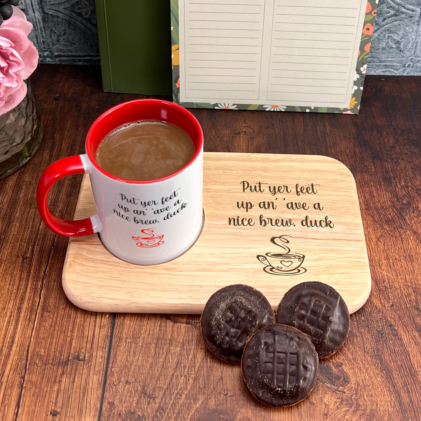 A wooden tray with a red and white coffee mug, a wooden cutting board with a message, and three chocolate-covered cookies on a wooden surface.