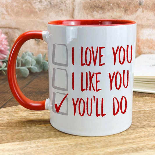 A white mug with a red handle, featuring a humorous text that reads "I LOVE YOU I LIKE YOU YOU'LL DO" along with checkboxes.