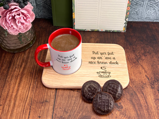 A wooden tray with a red and white coffee mug, a wooden cutting board with a message, and three chocolate-covered cookies on a wooden surface.
