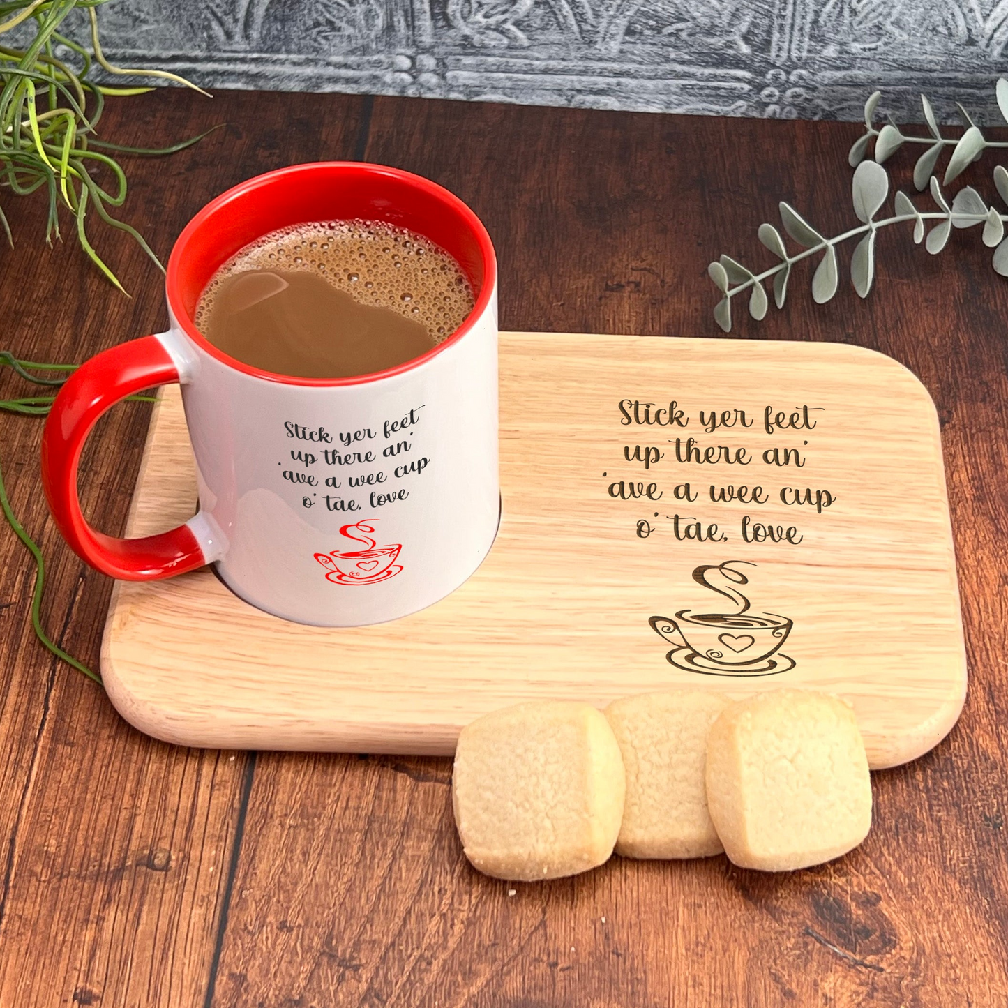 A red and white coffee mug with a wooden tray and cookies on a wooden surface, accompanied by a plant and a wooden board with a message.