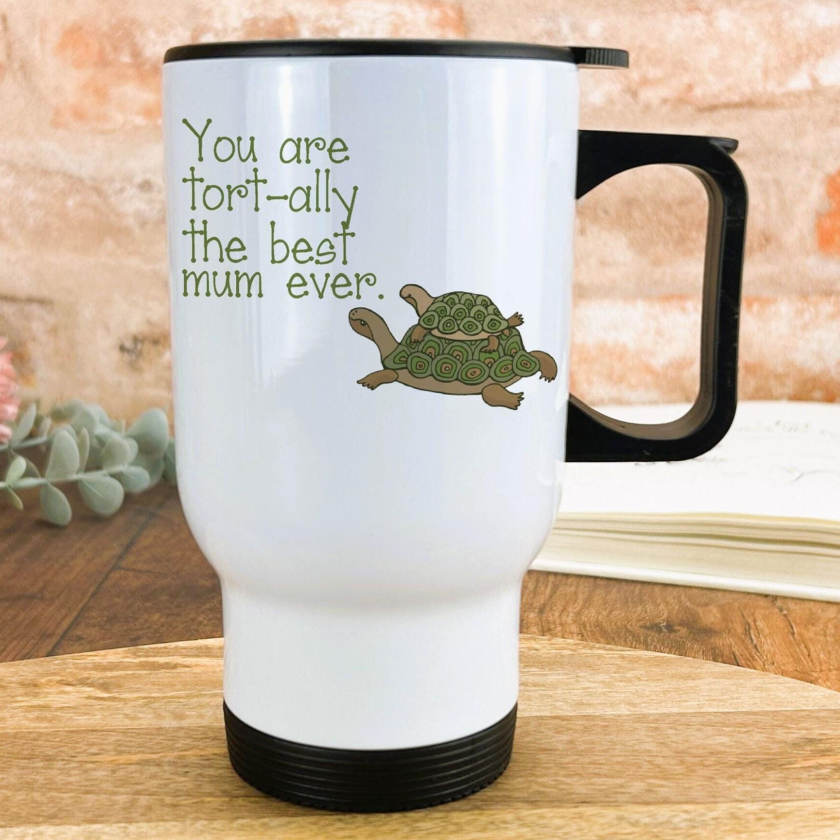 A white travel mug with a black handle, featuring a turtle design and text that reads "You are fort-ally the best mum ever."