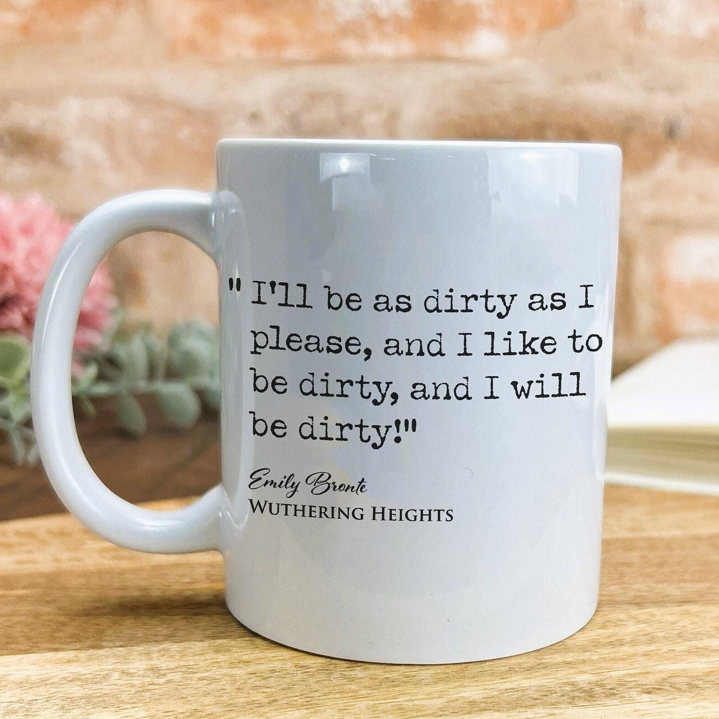 A white ceramic mug with a quote from the novel "Wuthering Heights" by Emily Brontë printed on it, placed on a wooden surface against a brick wall background.