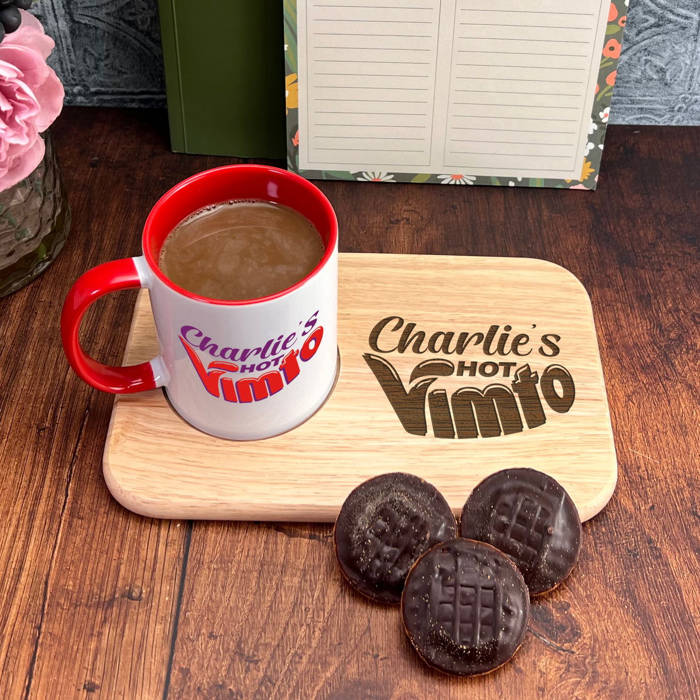 A mug of coffee and three chocolate-covered cookies are placed on a wooden tray, accompanied by a vase of pink flowers and a notebook with a floral design.