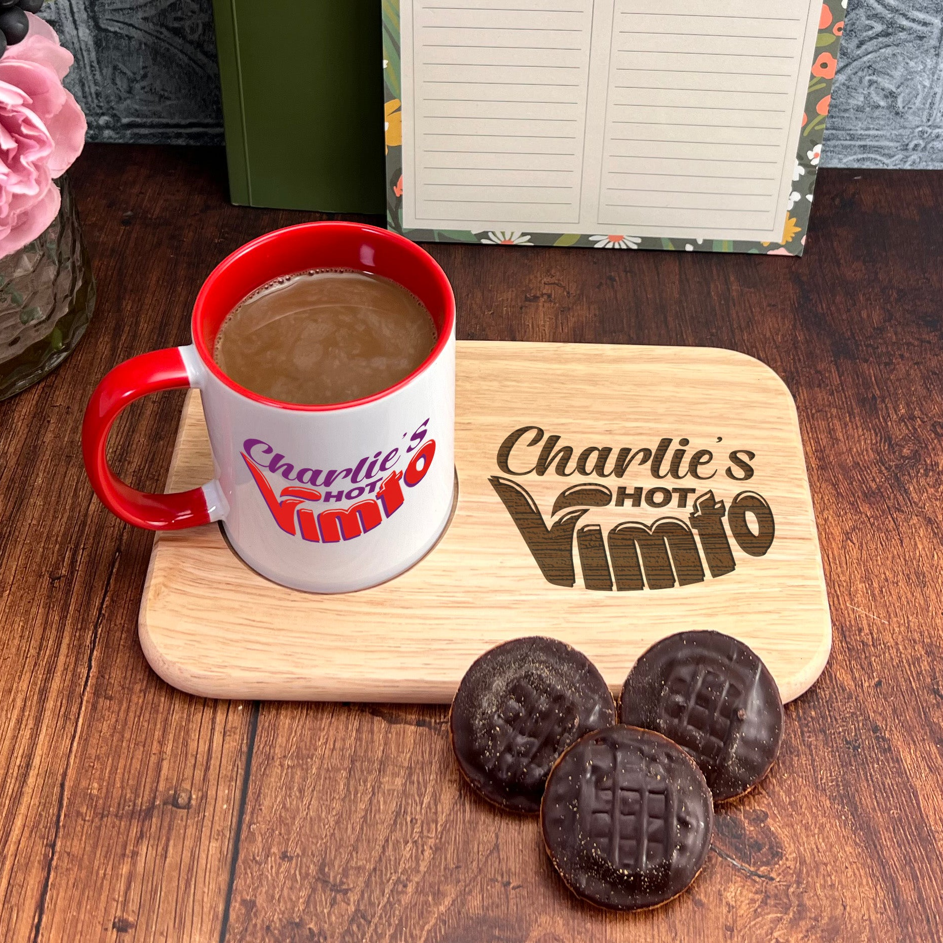A mug of coffee and three chocolate-covered cookies are placed on a wooden tray, accompanied by a vase of pink flowers and a notebook with a floral design.